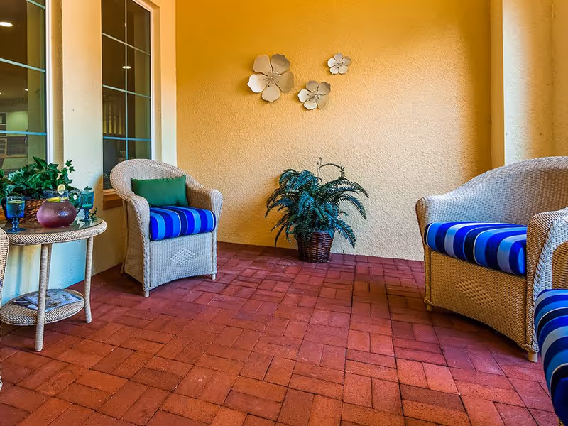 A cozy outdoor seating area with two wicker chairs and a wicker loveseat, all with blue and green striped cushions. There is a small wicker side table with a plant and decorative items on it. The floor is made of red brick tiles, and the walls are painted yellow with three white flower wall decorations. A potted green plant is placed against the wall.