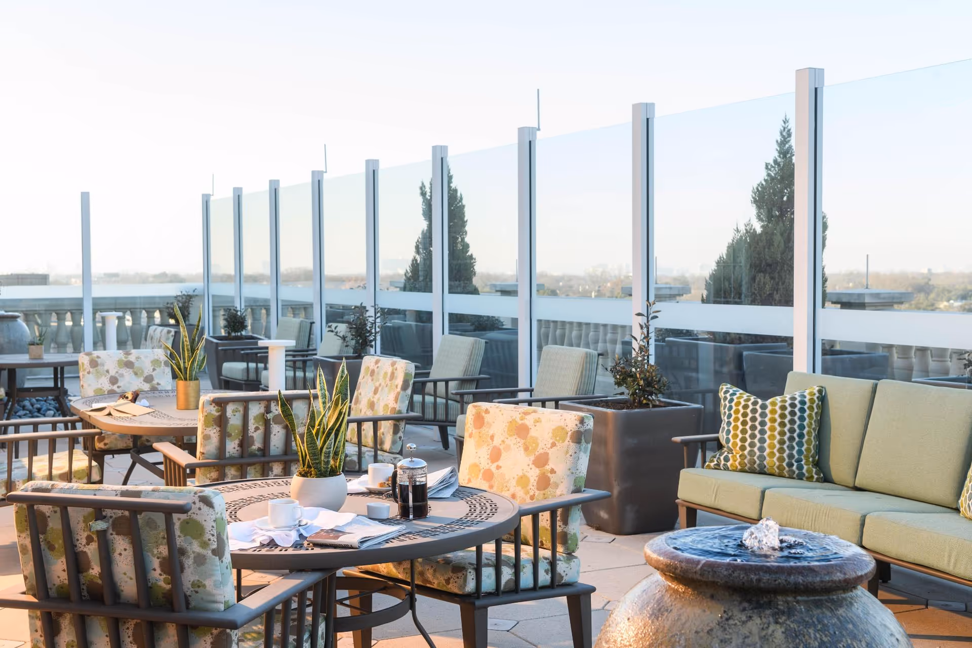 Outdoor rooftop terrace with round tables, cushioned chairs, potted plants, a sofa and a small fountain against a glass windscreen.