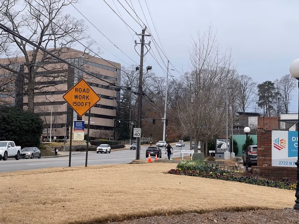 Street view showing a 'ROAD WORK 1500 FT' sign, cars and pedestrians with office buildings and trees in the background.