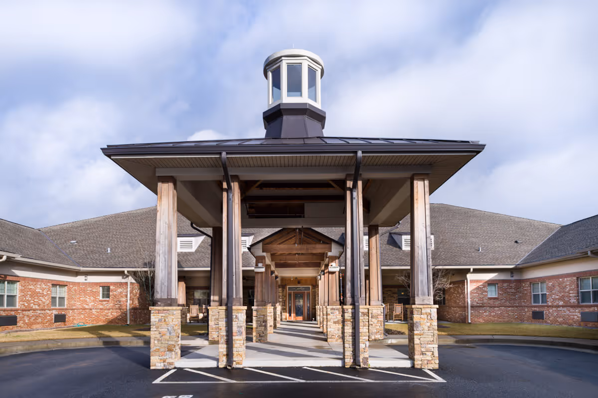 Front exterior view of Hope Memory Care Center showing a covered entrance with wooden pillars and stone bases, a peaked roof with a small cupola on top, and brick walls on either side under a partly cloudy sky.