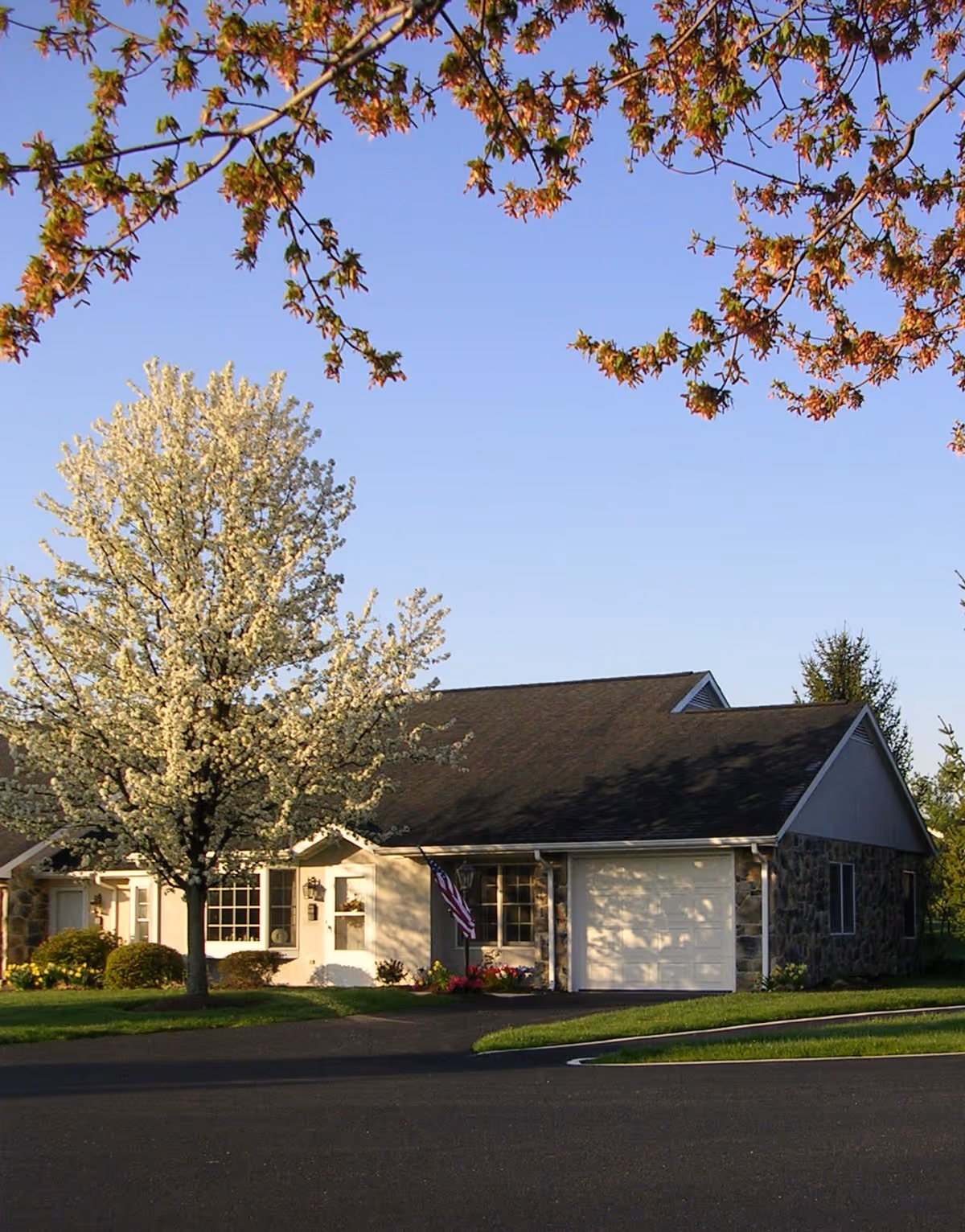 Single-story residential building with a stone and siding exterior, a garage, and an American flag near the entrance. The building is surrounded by a well-maintained lawn and blooming trees under a clear blue sky.