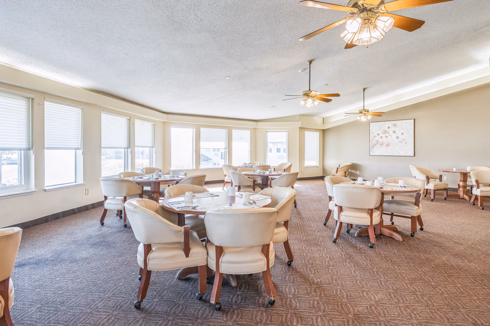 A bright dining room with multiple round tables surrounded by beige cushioned chairs on wheels. The room has large windows with white blinds, beige walls, carpeted floor, ceiling fans with lights, and a floral painting on one wall.