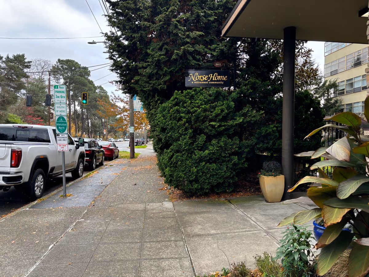 Sidewalk and street in front of Norse Home retirement community with a sign, parked cars, trees, and a partially visible building entrance.