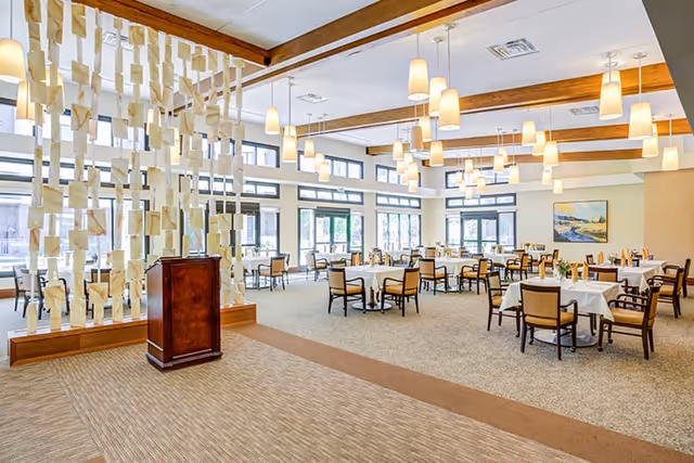 Spacious dining room with multiple round tables covered with white tablecloths and surrounded by wooden chairs with cushions. The room features large windows allowing natural light, modern pendant lighting hanging from a ceiling with wooden beams, and a decorative partition with hanging rectangular pieces near a wooden podium. A painting is visible on the far wall.