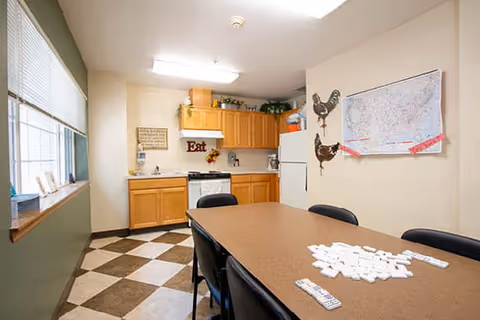 A kitchen and dining area in a senior living facility with wooden cabinets, a white refrigerator, a stove, and a table with chairs. The table has domino tiles and a remote control on it. The walls are decorated with a map and rooster wall hangings, and there is a window with blinds letting in natural light.