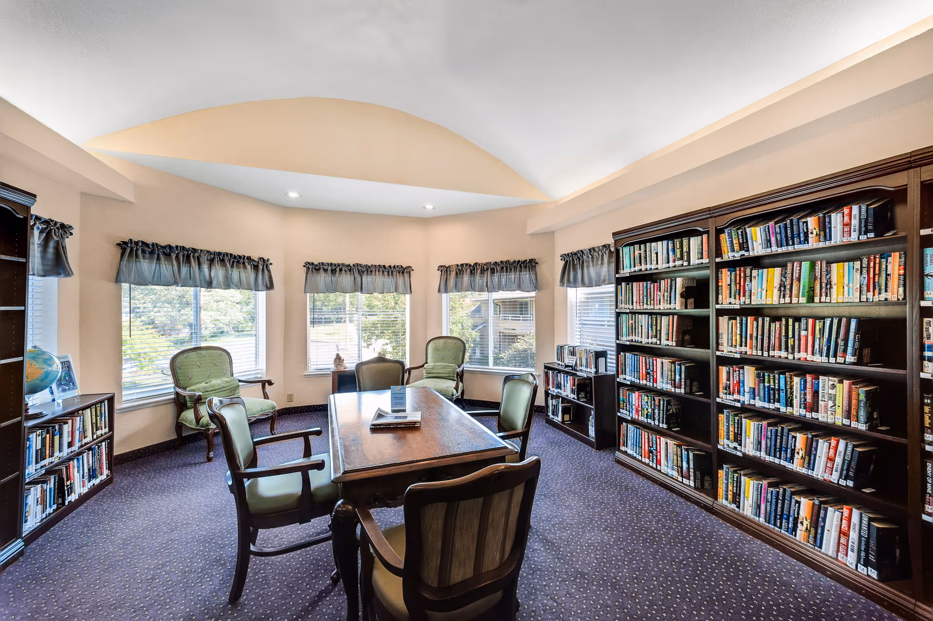 A cozy library room with large windows covered by short curtains, a wooden table surrounded by six chairs with green upholstery, and multiple bookshelves filled with books. The room has a purple carpet and a vaulted ceiling with recessed lighting.