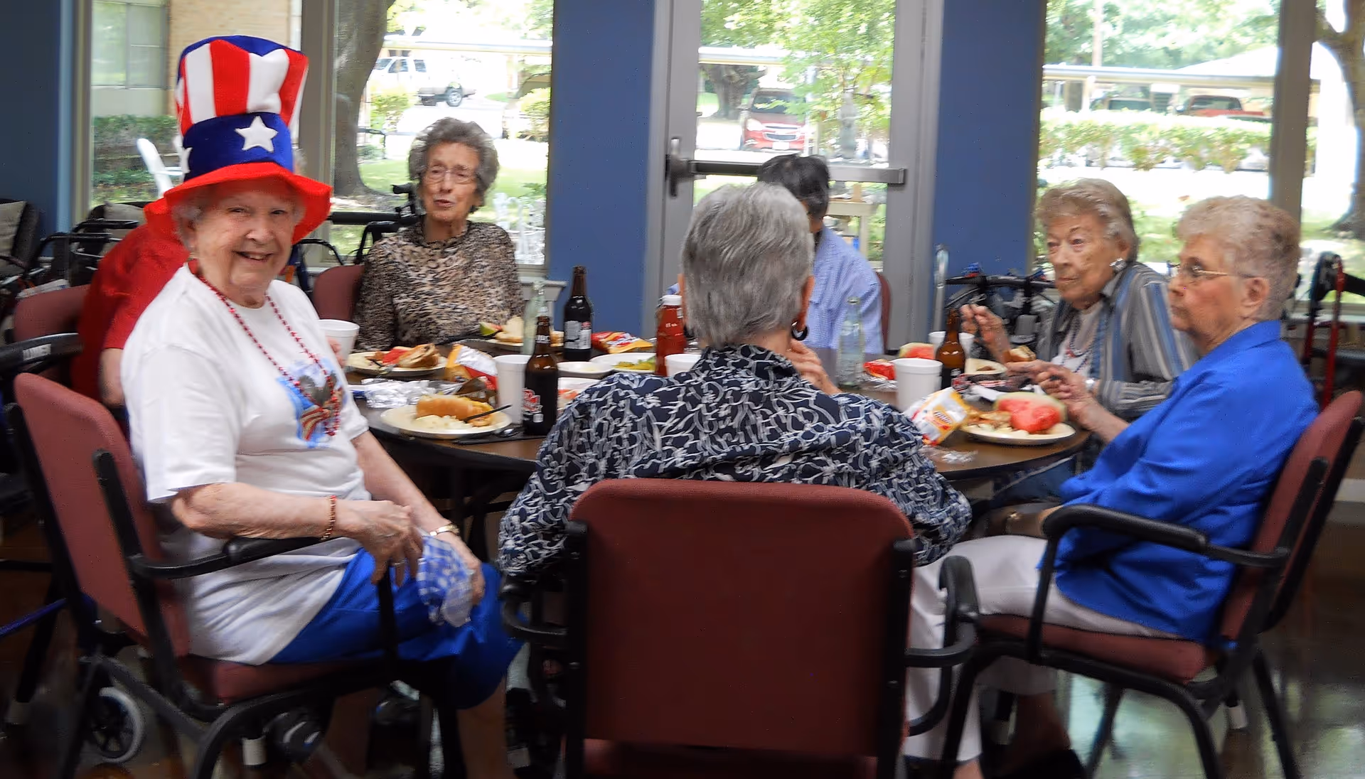 Several elderly women sit around a dining table eating and socializing, one smiling and wearing a tall red, white, and blue patriotic hat.
