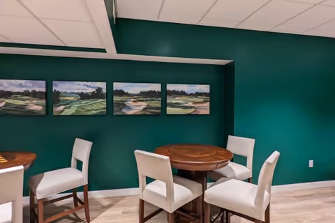 A room with dark green walls featuring four landscape paintings hung in a row. There are two round wooden tables, each surrounded by four white cushioned chairs with wooden legs. The floor is light-colored wood, and the ceiling has white tiles with recessed lighting.