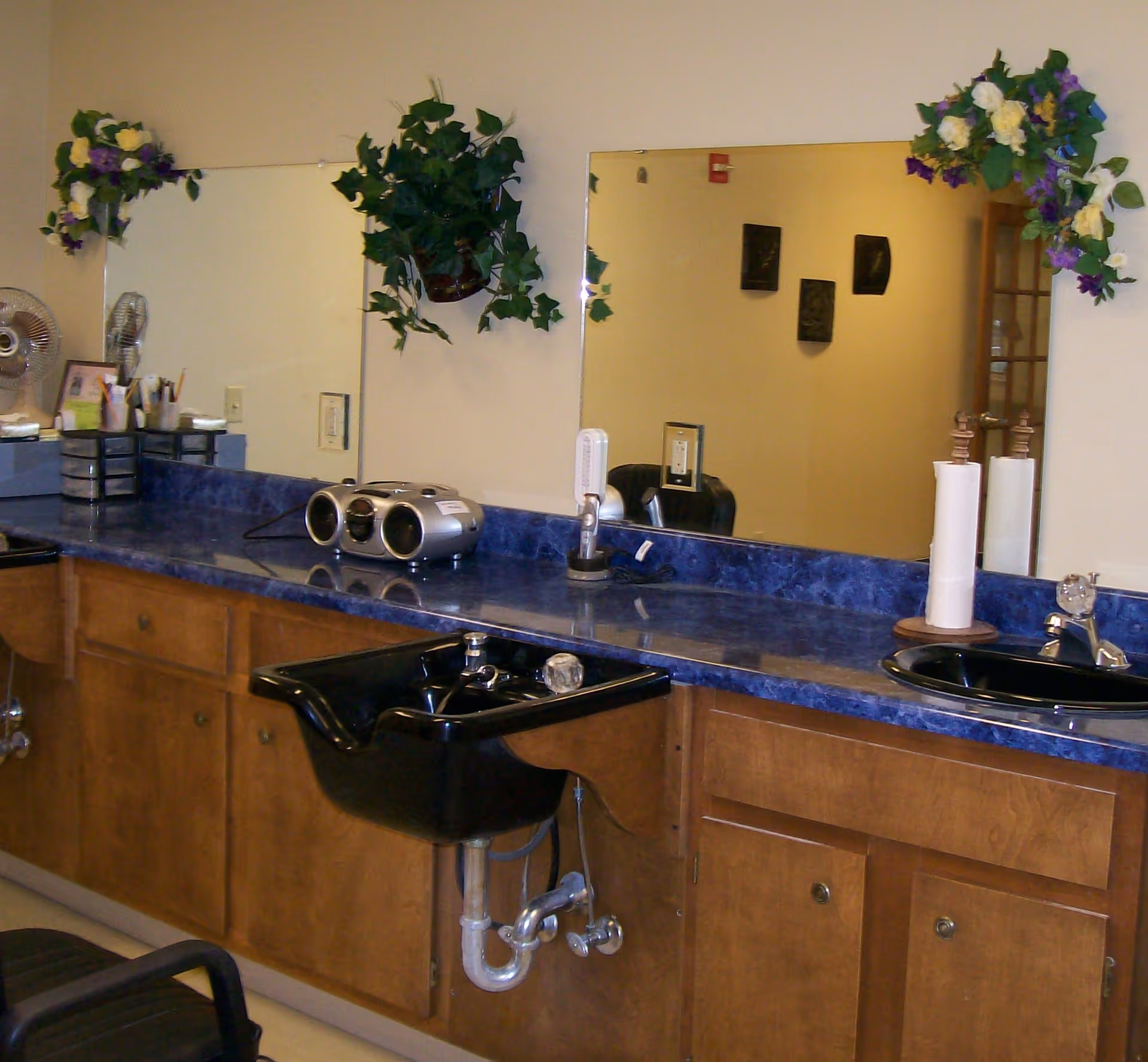 Interior view of a salon area with a blue countertop, two black sinks, large mirrors decorated with artificial flowers, a small stereo, a fan, and various salon supplies on the counter.