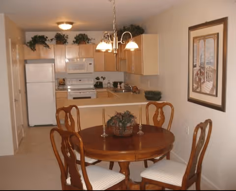A small dining area with a round wooden table and four chairs with white cushions. On the table is a decorative centerpiece and two candlesticks. Behind the dining area is a compact kitchen with light wood cabinets, a white refrigerator, microwave, stove, and sink. There are some green plants on top of the cabinets and a framed picture on the wall to the right.