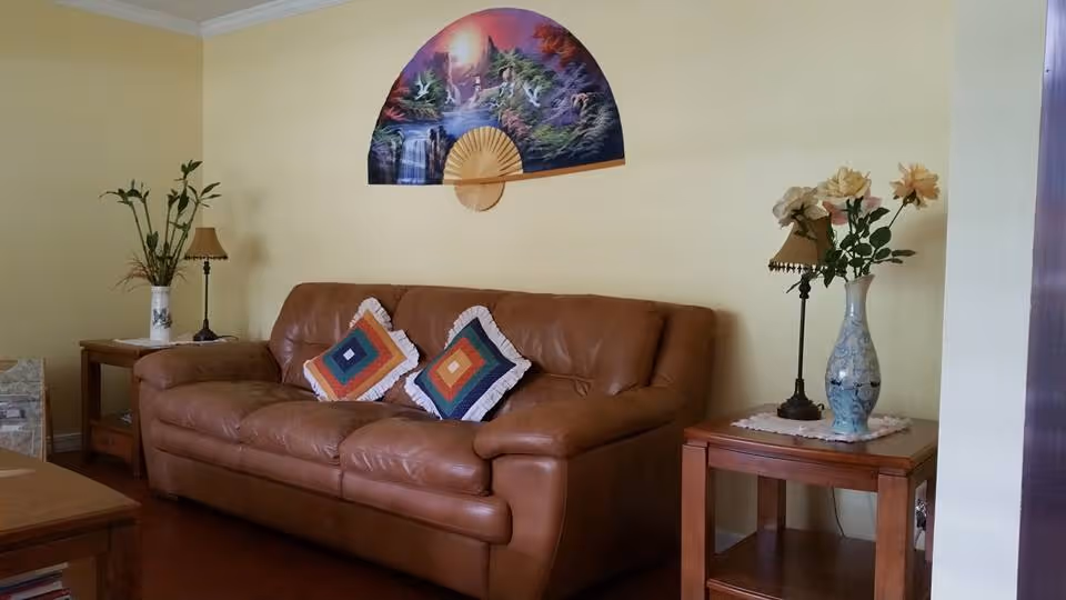 A cozy living room with a brown leather sofa adorned with two colorful square pillows. On either side of the sofa are wooden side tables, each holding a decorative lamp and a vase with flowers or plants. Above the sofa hangs a large decorative fan with a painted nature scene featuring birds, trees, and a waterfall. The walls are painted light yellow, and the floor is wooden.