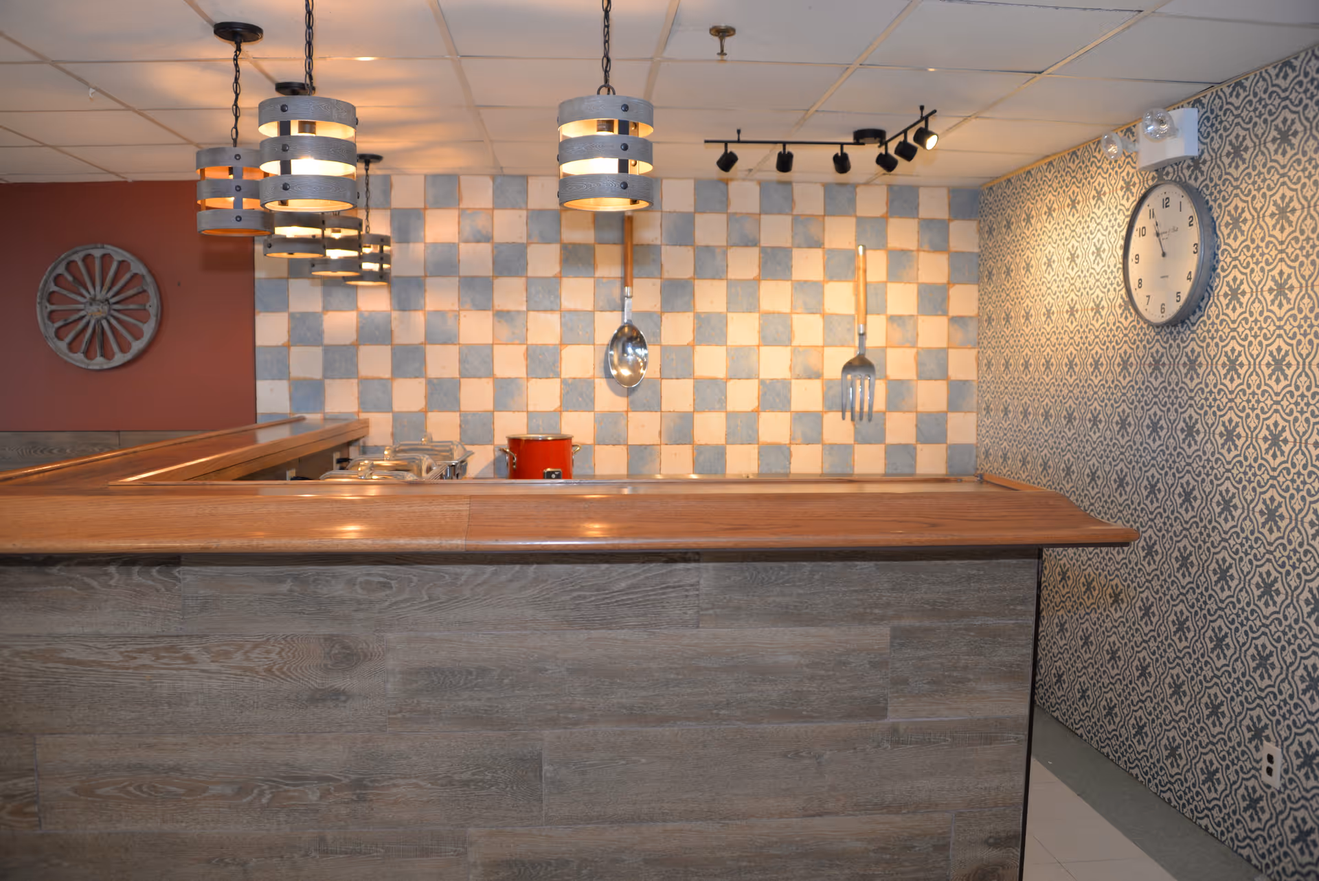 Interior view of a kitchen area with a wooden counter in the foreground, hanging pendant lights above, a checkered tile backsplash, and large decorative utensils hanging on the wall. A clock is mounted on a patterned wall to the right.