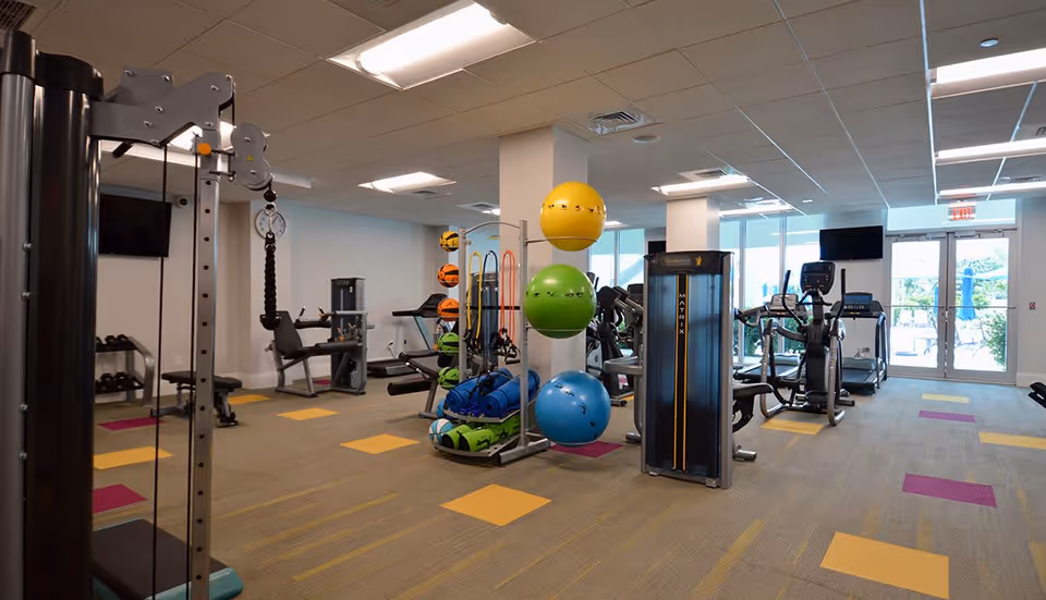 A well-lit fitness room with various exercise equipment including weight machines, treadmills, stationary bikes, and colorful exercise balls. The room has large windows and glass doors leading to an outdoor area.