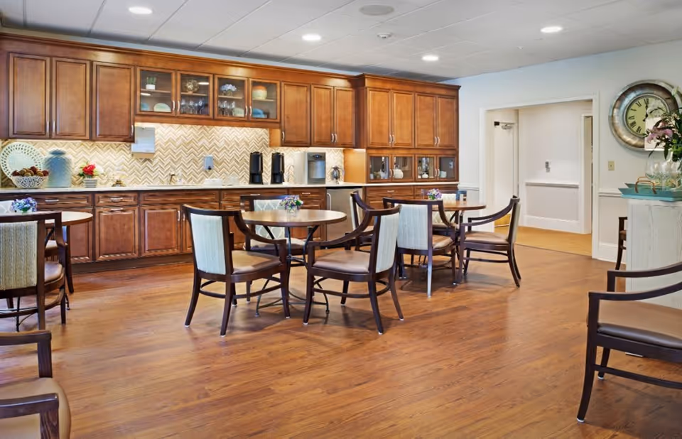 A dining area with several round tables and chairs on a wooden floor. The back wall features wooden cabinets with glass doors, a countertop with a coffee machine, and decorative items. A large clock hangs on the right wall above a small cabinet with flowers and glassware.