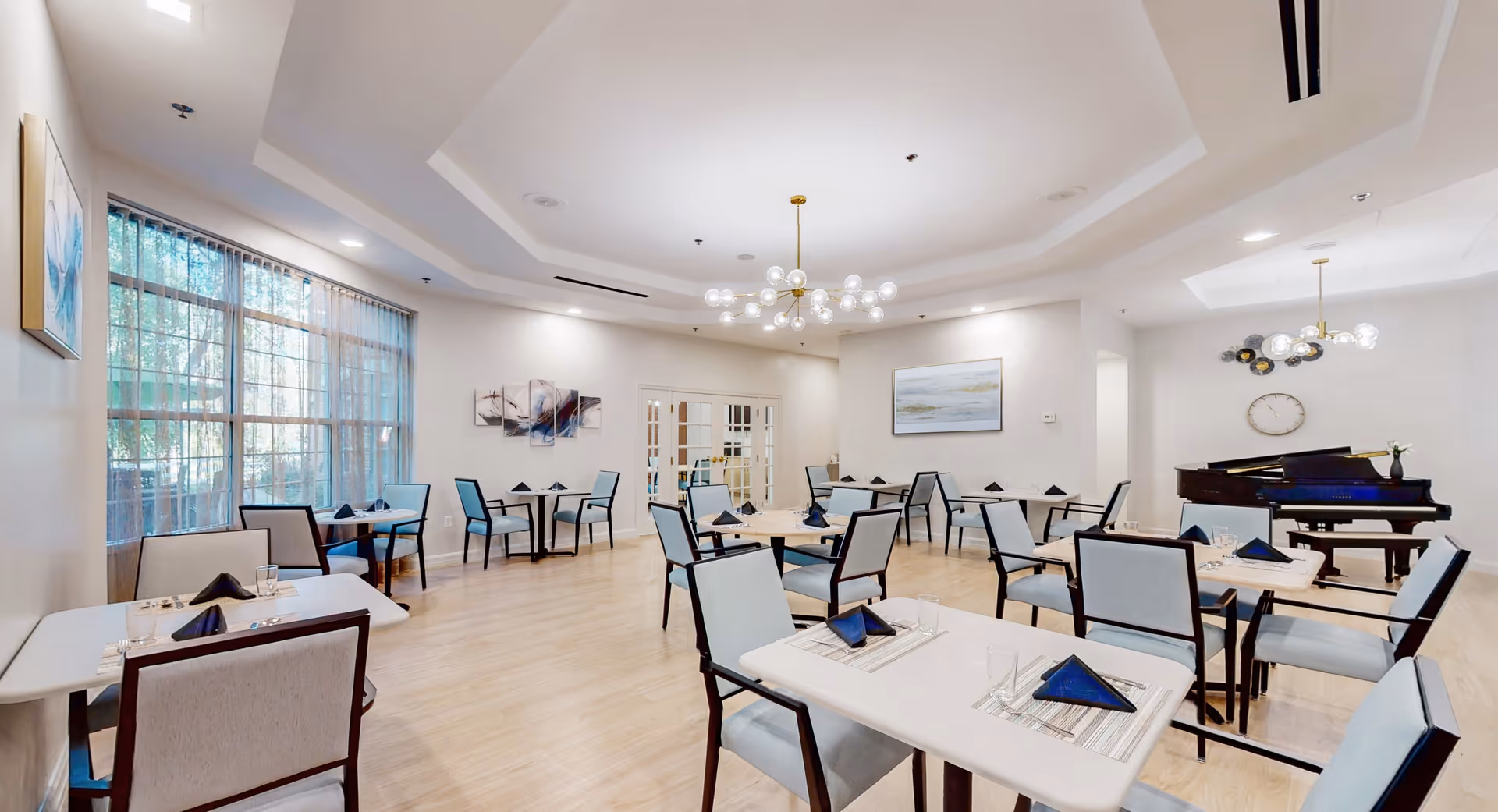 A bright and spacious dining room with multiple tables and chairs arranged neatly. Each table is set with placemats, glasses, and folded dark blue napkins. Large windows with sheer curtains allow natural light to fill the room. The walls are decorated with modern artwork, and a grand piano is positioned in the corner under a contemporary chandelier. The flooring is light wood, and the ceiling features recessed lighting and a stylish central light fixture.