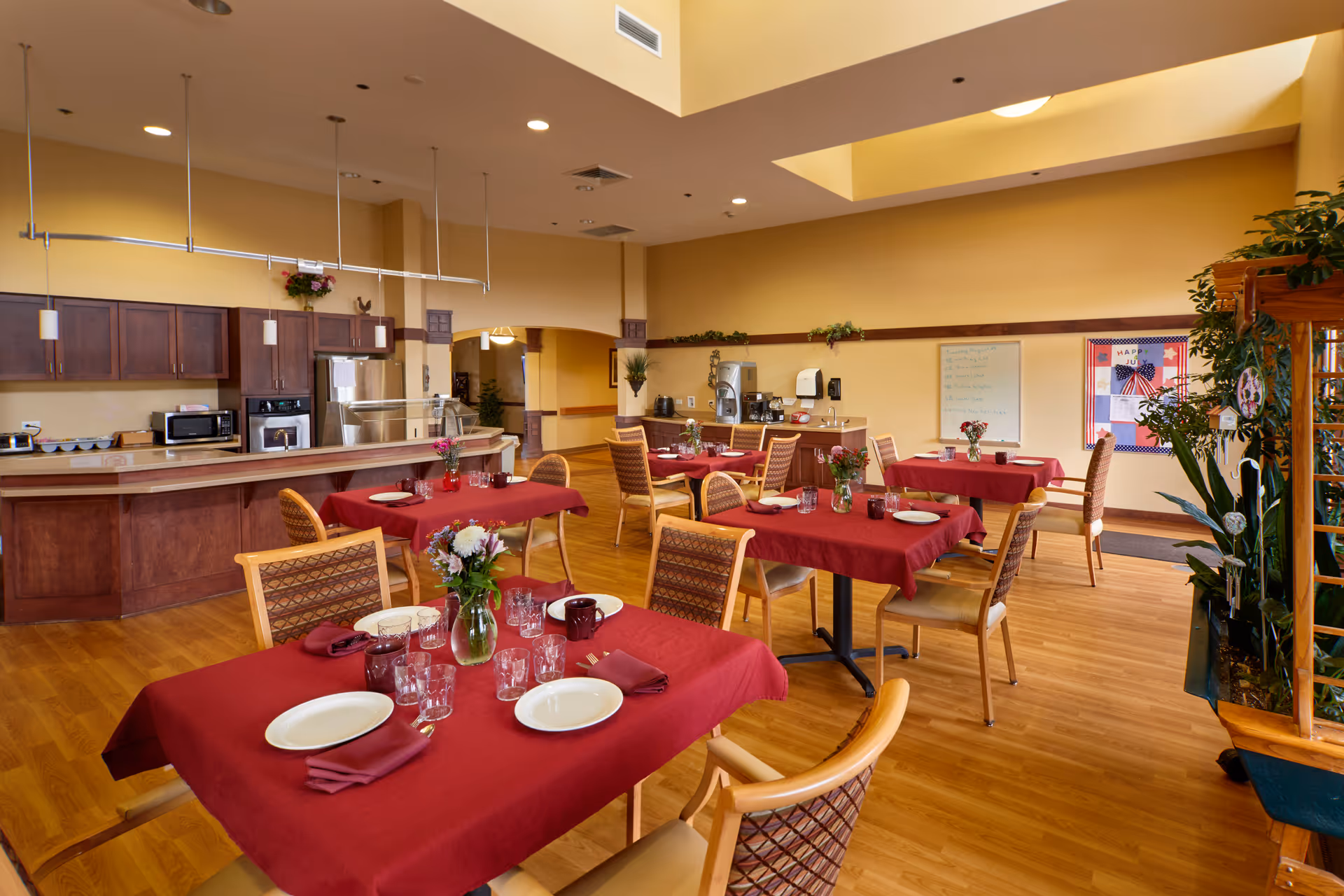 A dining room in a senior living facility with several tables covered in red tablecloths, each set with plates, glasses, and napkins. The room features wooden flooring, a kitchen area with cabinets and appliances in the background, and some plants and decorations around the space.