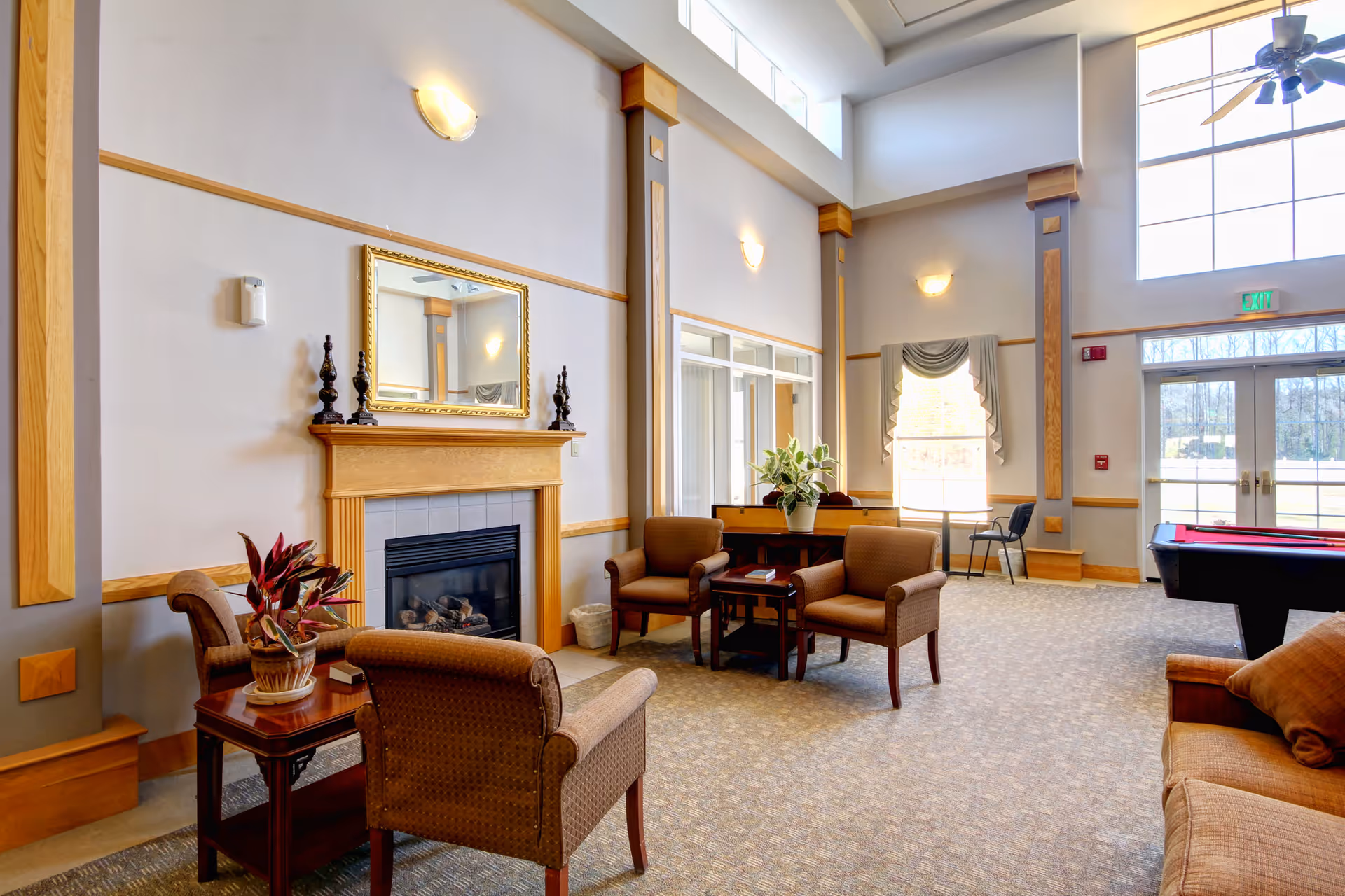 Bright communal lounge with high ceilings, a fireplace, several armchairs and tables, plants, and a pool table near large windows.