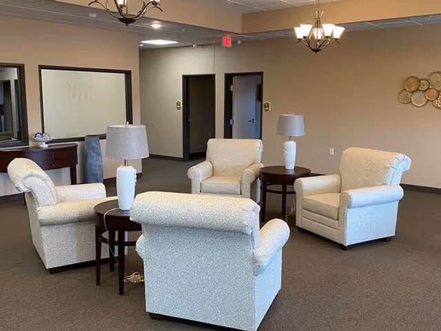 A seating area in an assisted living facility with four cushioned armchairs arranged around two small round wooden tables, each table holding a white lamp. The room has beige walls, a carpeted floor, and a decorative wall piece with circular elements. In the background, there are two open doorways and a console table with a mirror above it.