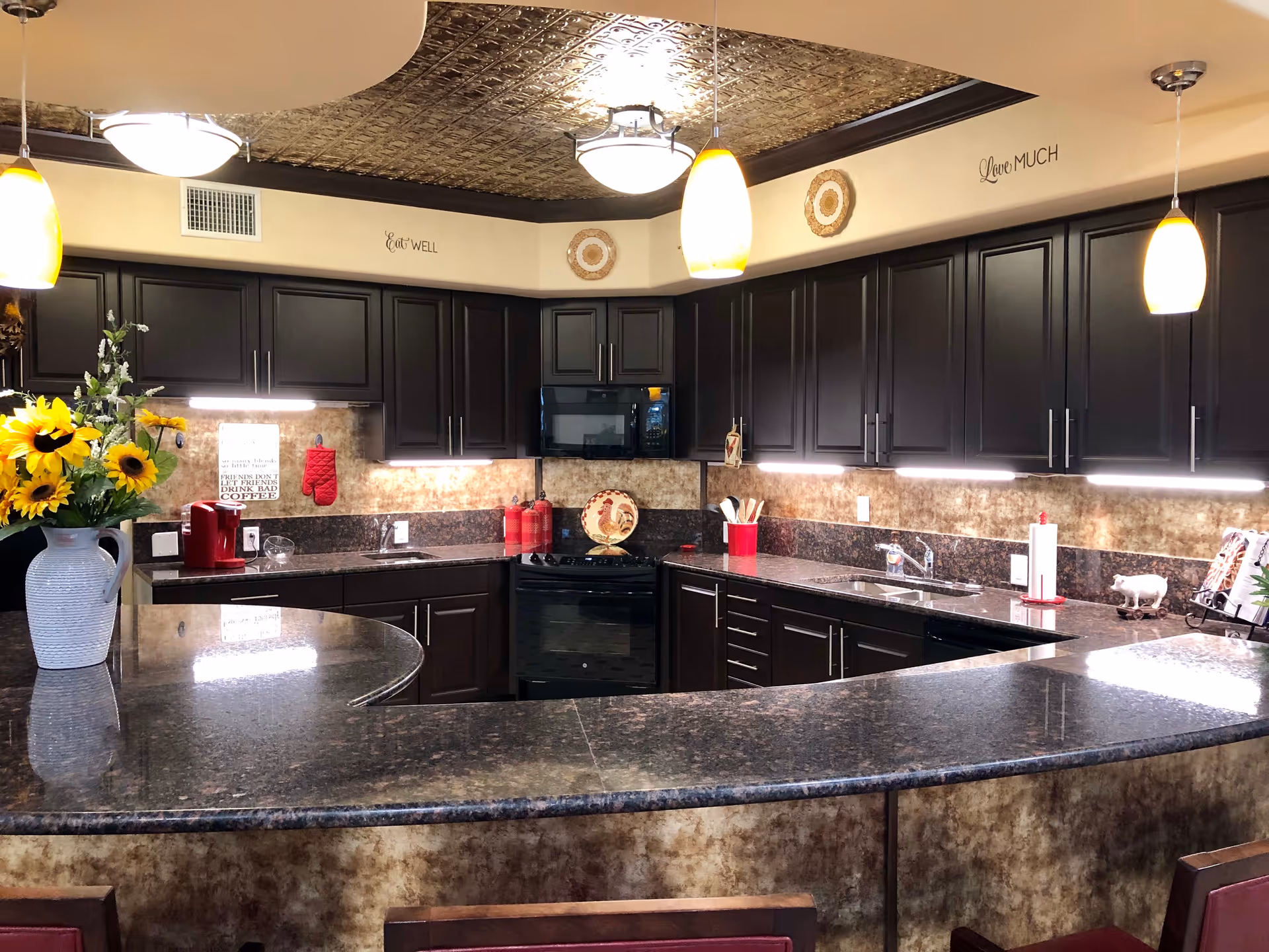 A spacious kitchen with dark wood cabinets and granite countertops. The kitchen features a black stove, microwave, and two sinks. There are three pendant lights hanging from the ceiling, which has a decorative metallic tile design. A vase with sunflowers is placed on the counter, and various kitchen utensils and decorations are visible. On the wall above the cabinets, there are decorative plates and the phrases 'Eat Well' and 'Love Much'.