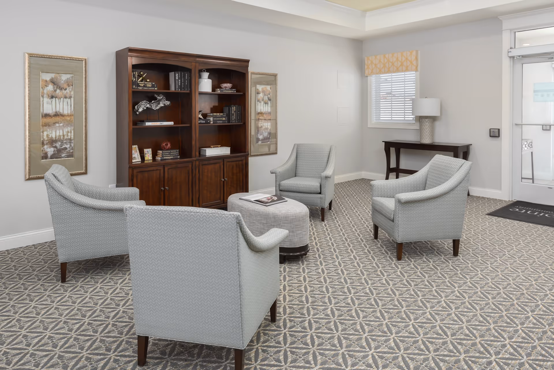 A cozy sitting area with four light gray upholstered armchairs arranged around a small oval ottoman. Behind the chairs is a dark wooden bookshelf filled with books and decorative items. The room has patterned carpet, light gray walls, two framed landscape paintings, a window with blinds and a yellow valance, a small wooden console table with a lamp, and a glass door leading outside.