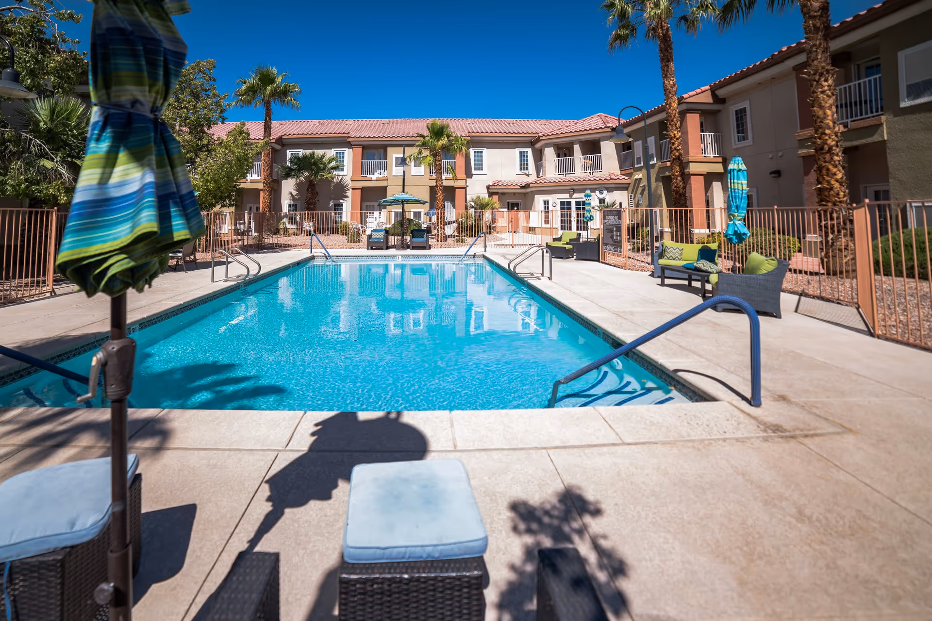 Outdoor swimming pool area at Acacia Springs facility with clear blue water, surrounded by lounge chairs, umbrellas, palm trees, and a two-story building in the background under a clear blue sky.
