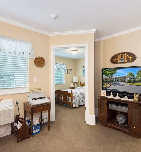 Interior view of a living area in an assisted living facility showing a small wooden table with a typewriter and a magazine rack near a window with blinds and a white valance. A doorway leads to a bedroom with a bed covered in floral bedding, a nightstand with a lamp, and framed pictures on the walls. A wooden TV stand holds a flat-screen television displaying an exterior image of the facility, along with decorative items and a DVD player.