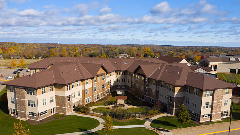 Aerial view of The Fountains at Hosanna senior living facility showing a large, U-shaped three-story building with brown roofs and beige walls. The building surrounds a landscaped courtyard with pathways, greenery, and a small gazebo. The surrounding area includes parking lots, trees with autumn foliage, and a partly cloudy sky.