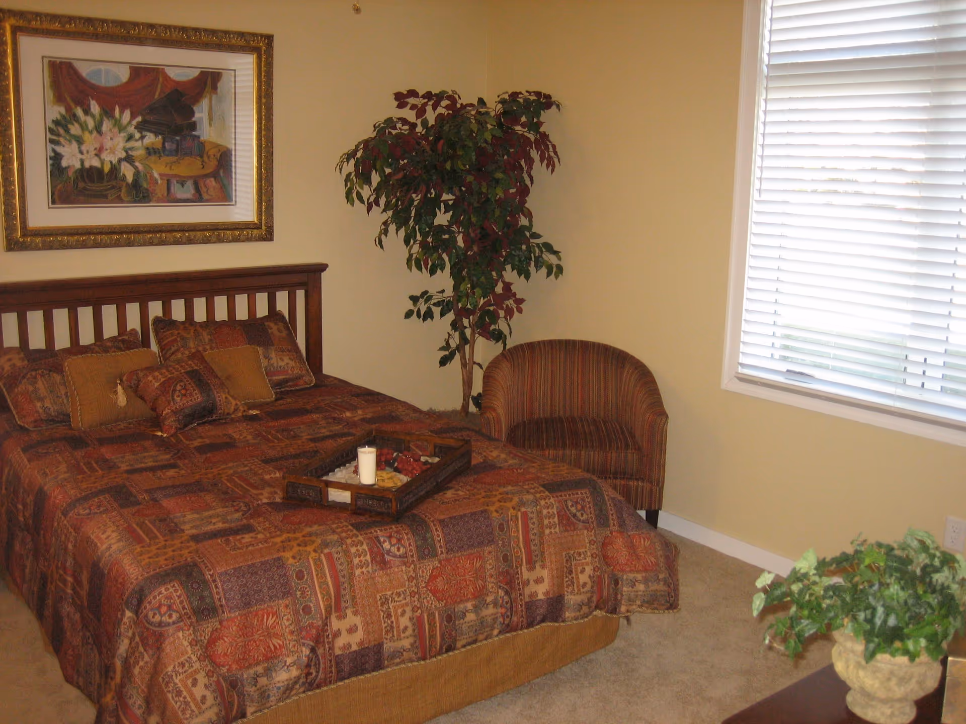 A bedroom with a wooden bed frame and a bed covered in a patterned quilt with multiple pillows. There is a tray with a candle and decorative items on the bed. A striped armchair is placed near a window with closed blinds. A large potted plant is in the corner, and a framed painting hangs on the wall above the bed. A small green plant is on a piece of furniture in the foreground.