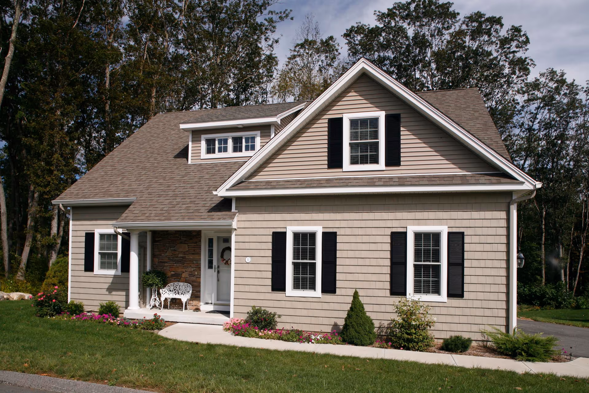 A beige single-family house with black shutters and a gray shingled roof, surrounded by green grass and trees. The house has a small porch with a white bench and some potted plants, and a concrete walkway leading to the front door.