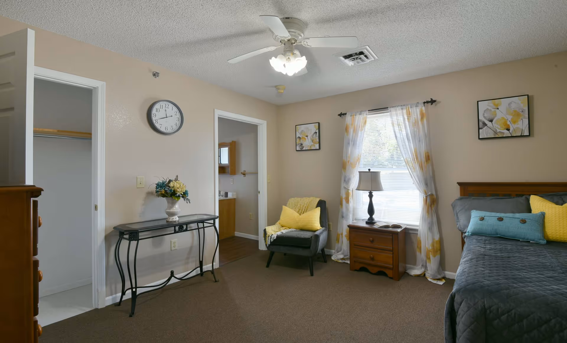 A bedroom with a bed on the right side covered with a gray quilt and decorative pillows in yellow and blue. There is a wooden nightstand with a lamp and an open book in front of a window with white and yellow floral curtains. A gray armchair with a yellow pillow and throw blanket is next to the nightstand. On the left wall, there is a black metal console table with a flower vase underneath a round wall clock. Two doorways are visible, one leading to a closet and the other to a bathroom with wooden cabinets.