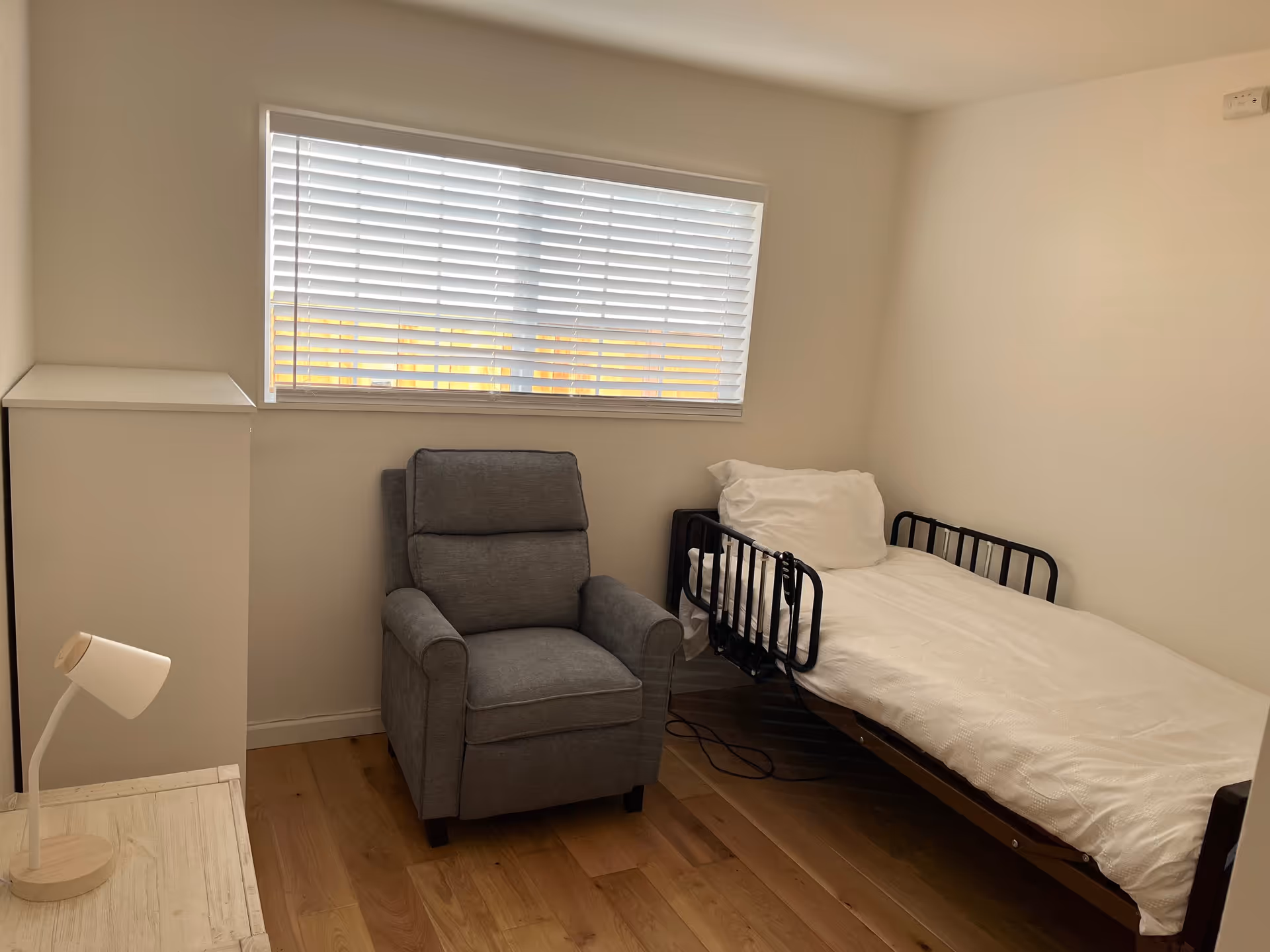 A small, simple bedroom with a single bed featuring black metal side rails, a white pillow and blanket, a gray upholstered armchair, a white cabinet, a wooden nightstand with a small white lamp, and a window with white blinds letting in natural light.
