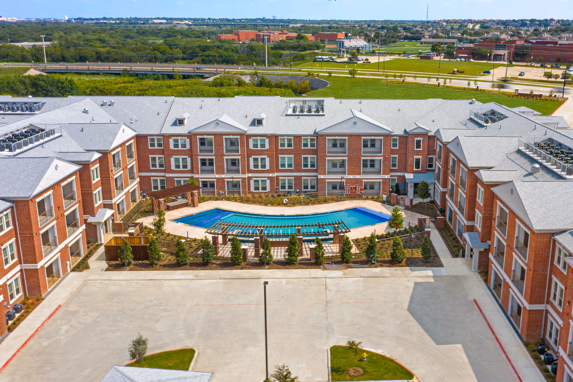 Aerial view of Presidium at Edgestone senior living facility showing a large U-shaped brick building surrounding a central outdoor swimming pool with pergolas and landscaped greenery. The background includes roads, parking areas, and distant buildings under a clear sky.