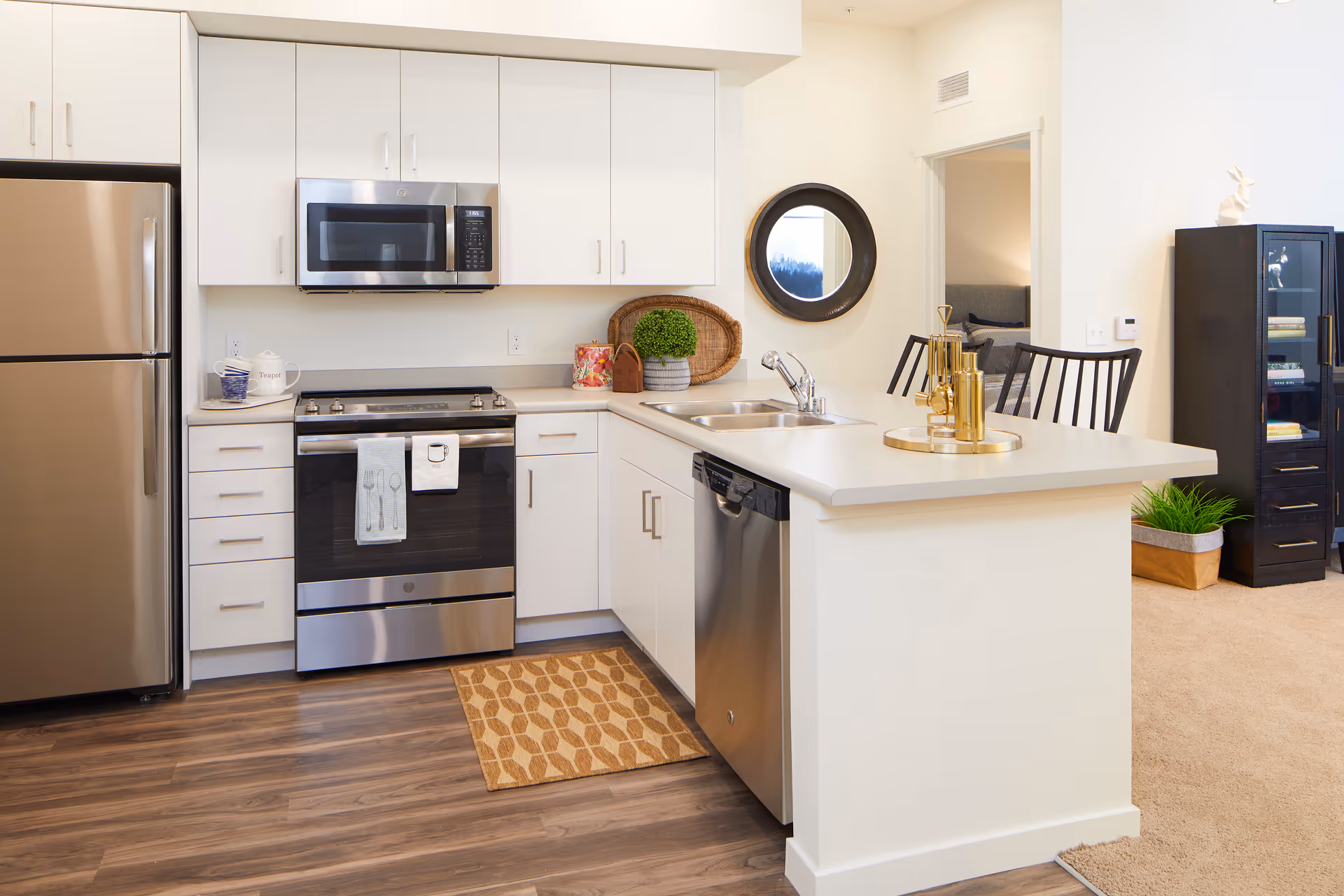 Modern kitchen with white cabinets, stainless steel refrigerator, oven, microwave, and dishwasher. The kitchen has a white countertop with a sink and gold decorative items on a tray. There is a small patterned rug on the wooden floor in front of the oven. A round mirror hangs on the wall near an open doorway leading to another room with a bed. A black cabinet with books and decorative items is visible in the adjacent living area.