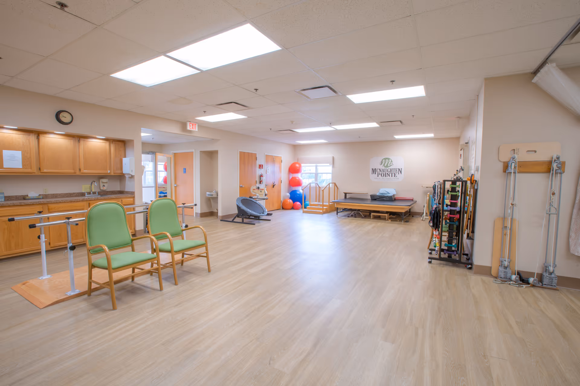 A spacious therapy or rehabilitation room with wooden flooring and beige walls. The room contains two green chairs, parallel bars for walking exercises, exercise balls stacked in the corner, a therapy table, and various physical therapy equipment along the walls. There is a sign on the far wall that reads 'McNaughten Pointe'.