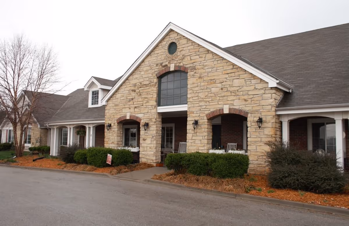 Exterior view of a single-story assisted living facility building with stone and brick facade, featuring a peaked roof, large windows, and a covered porch area with chairs. Shrubs and mulch landscaping surround the building, and a paved driveway is visible in front.