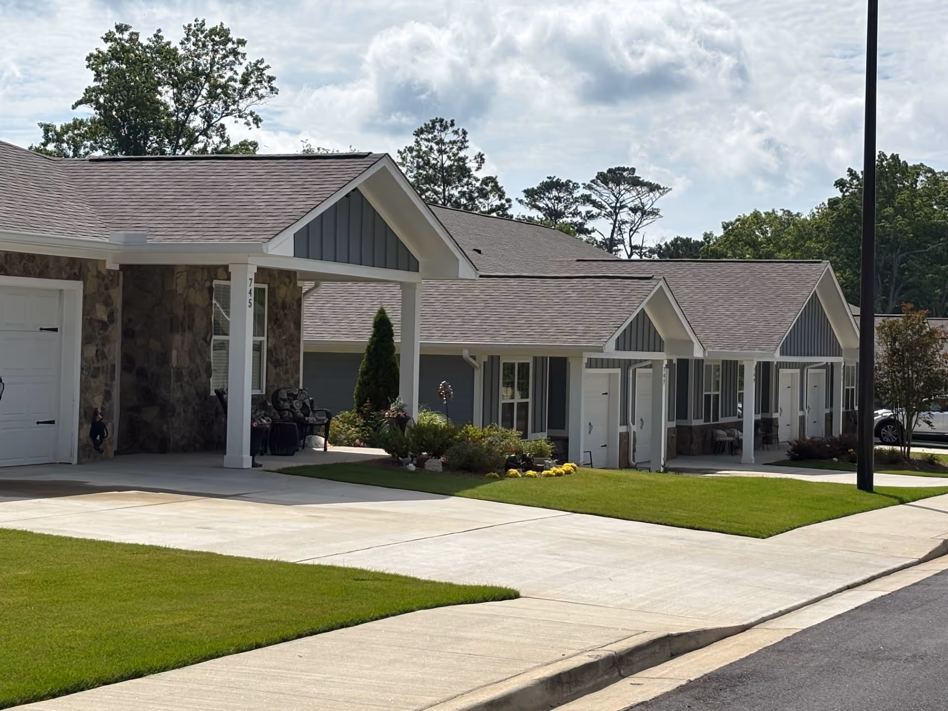 Row of single-story assisted living units with covered porches, manicured lawns, and a driveway under a partly cloudy sky.