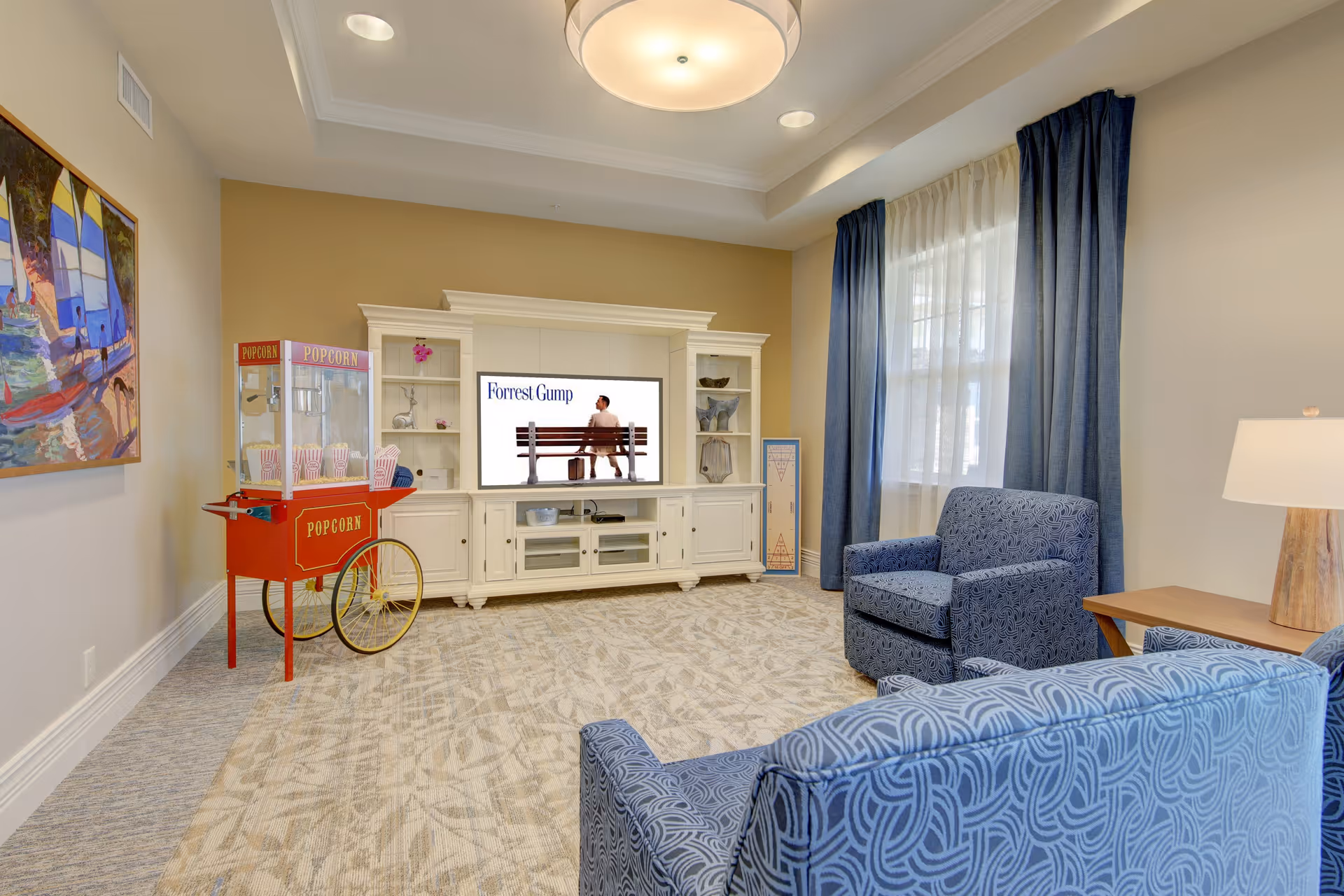 A cozy living room with two blue patterned armchairs and a matching sofa facing a white entertainment center with a TV displaying the movie Forrest Gump. A red popcorn machine with popcorn bags is positioned to the left, and a colorful painting of sailboats hangs on the wall. The room has beige walls, a patterned carpet, blue curtains, and a table lamp on a wooden side table.