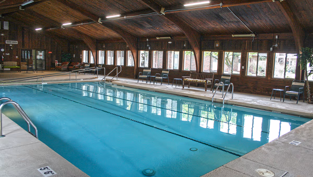 Indoor swimming pool in a wooden vaulted room with chairs and windows along the side.