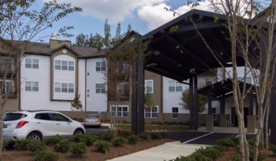 Exterior view of a senior living facility with a covered entrance driveway, landscaped bushes, trees, and parked cars in front of a multi-story building with white and beige siding.