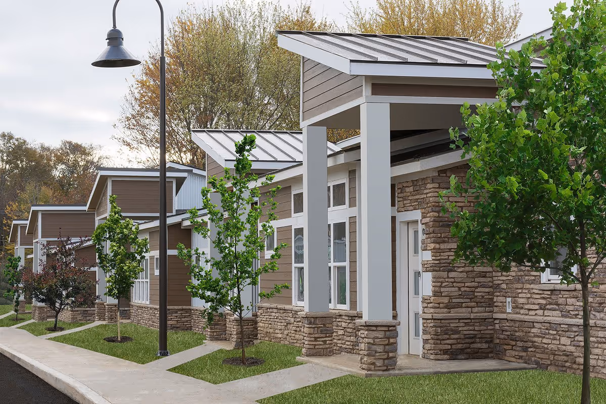 Exterior view of a modern senior living facility building with stone and wood siding, multiple windows, small trees planted along a sidewalk, and a street lamp.