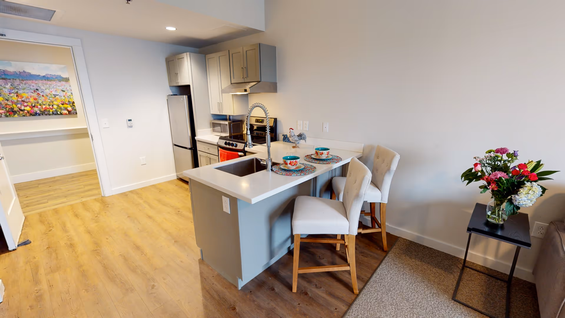 Modern kitchen area with light wood flooring, gray cabinets, stainless steel refrigerator and stove, and a white countertop island with a sink. Two cushioned chairs are placed at the island, set with colorful bowls and placemats. A small side table with a vase of flowers is next to a sofa on the right. A colorful painting is visible on the wall in the adjacent room.