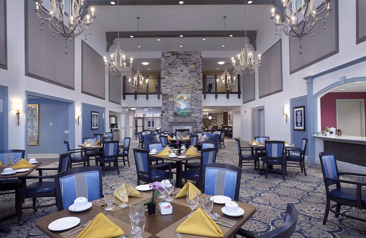 A spacious dining room with multiple tables set with yellow napkins, white plates, cups, and glassware. The room features blue cushioned chairs, a patterned carpet, and a large stone fireplace in the center background. Elegant chandeliers hang from the high ceiling, and there are decorative wall panels and framed artwork on the walls.