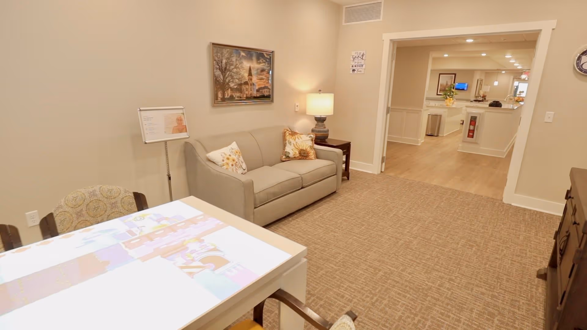 A cozy interior room with a beige couch adorned with floral pillows, a side table with a lamp, a framed picture on the wall, and a table with chairs. The room opens into a hallway with wooden flooring and bright lighting.