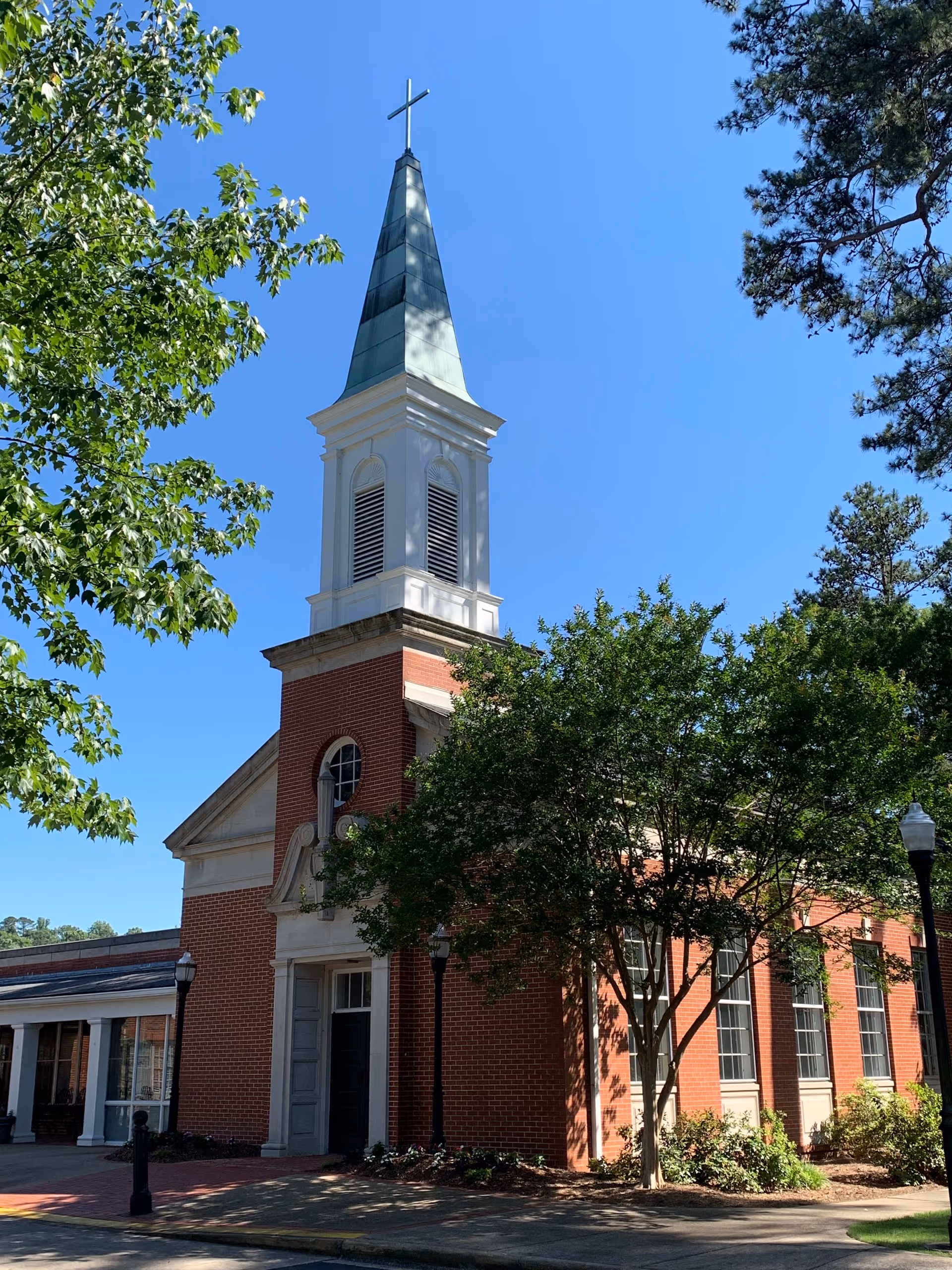 Exterior view of a red brick building with a tall white steeple topped with a cross, surrounded by green trees under a clear blue sky.