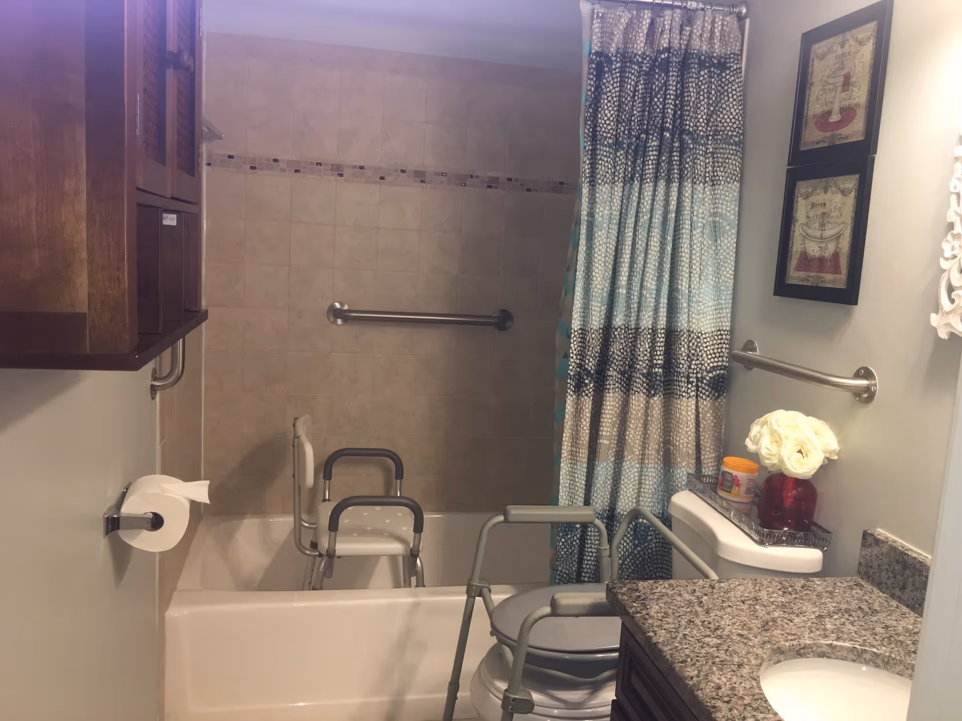 A bathroom with a bathtub and shower area featuring a grab bar and a shower chair. There is a toilet with safety rails around it, a granite countertop with a sink, a toilet paper holder, a wooden cabinet mounted on the wall, and a shower curtain with a blue and beige pattern. Decorative framed pictures and a vase with white flowers are placed on the toilet tank.