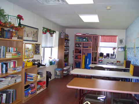 A bright activity room with folding tables and chairs, bookshelves, and storage cabinets along the walls.