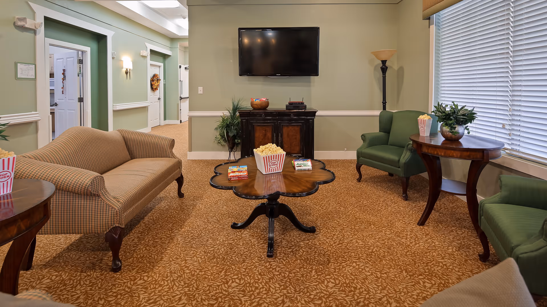 A cozy living room area in an assisted living facility with a patterned carpet, a beige sofa, two green armchairs, and wooden side tables. A wooden coffee table in the center holds popcorn and snacks. A flat-screen TV is mounted on the wall above a dark wooden cabinet. A tall floor lamp stands in the corner near a window with closed blinds.
