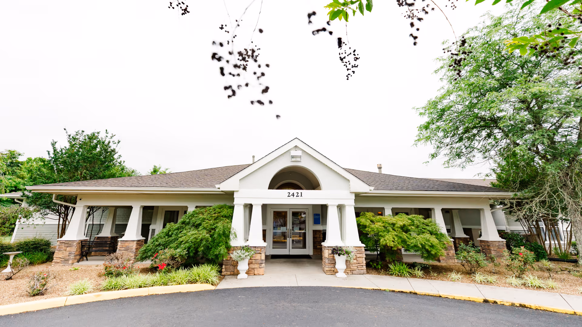 Front exterior view of a single-story building with white columns and stone bases, surrounded by greenery and trees. The building has a central entrance with double glass doors and the number 2421 displayed above. There is a curved driveway in front of the entrance.