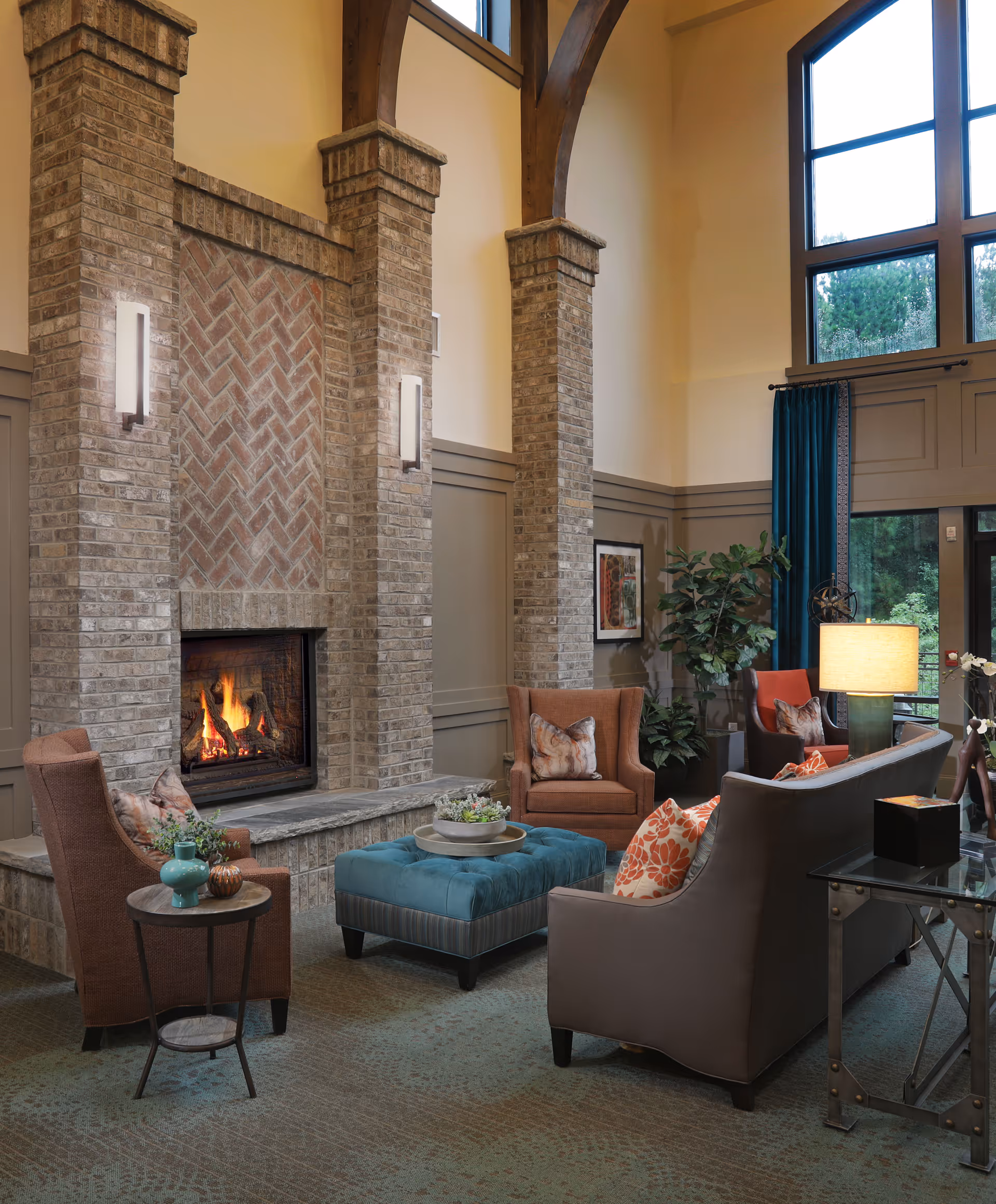 A cozy living room area with a large brick fireplace, two brown armchairs with decorative pillows, a gray sofa with orange and white pillows, a blue tufted ottoman, a side table with decorative items, a floor lamp, and large windows with blue curtains letting in natural light.