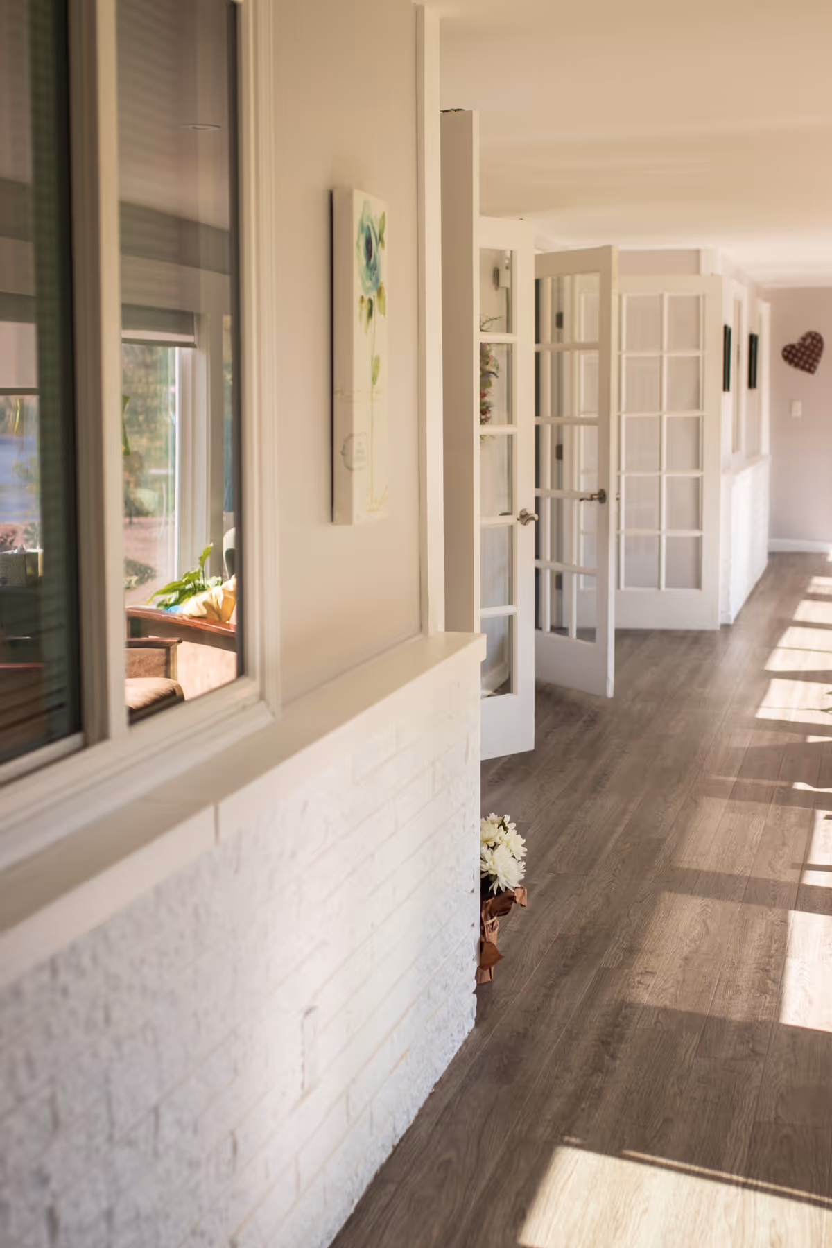 Sunny interior hallway with wood floors, a white half-brick wall, windows and open French doors.